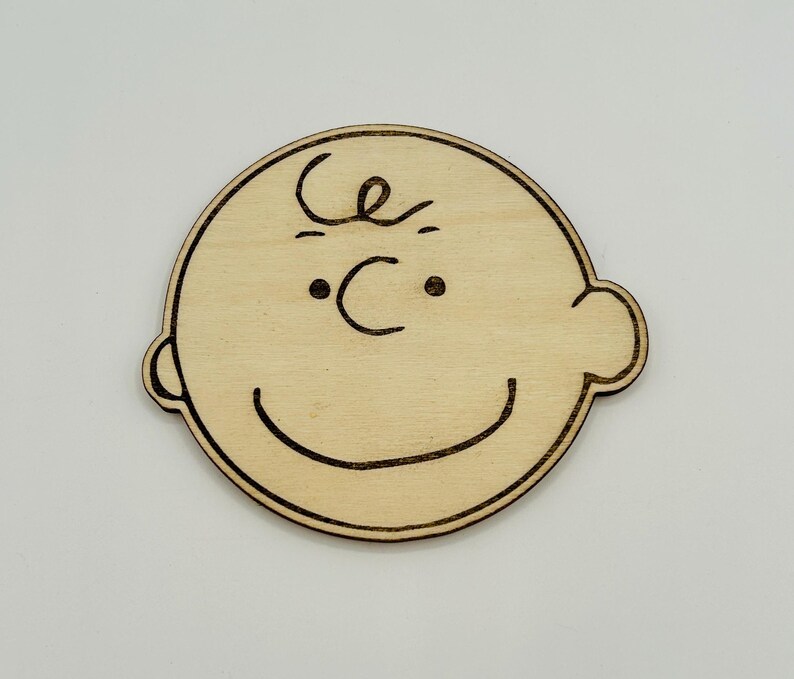 May include: A wooden cutout of a cartoon character's face. The design features a round head, small eyes, a curved smile, and a single curl of hair. The character's features are outlined in dark brown against the light wood.