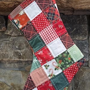 May include: A quilted Christmas stocking with a patchwork design. The stocking features squares of red, green, and white fabric with patterns like plaid, polka dots, and holiday motifs. A red and white gingham loop hangs from the top.