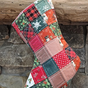 May include: A quilted Christmas stocking with a patchwork design. The stocking features squares of red, green, and white fabrics with holiday patterns like snowflakes, plaid, and holly. A red and white polka dot loop hangs from the top.