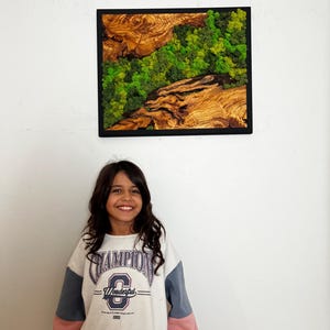 May include: Square wall art with a black frame. The artwork features a natural landscape design with wood and green moss. The wood has a rich, brown color, and the moss is a vibrant green. The artwork is hanging on a white wall.
