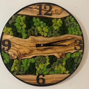 Puede incluir: Un reloj redondo con marco negro y números romanos. La esfera del reloj está hecha de madera con veta de madera natural y detalles de musgo verde.