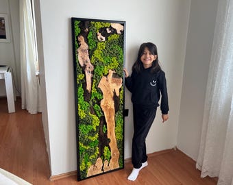 Può includere: Un'opera d'arte da parete incorniciata in nero che presenta un tronco d'albero conservato con muschio verde che cresce su di esso. Una persona giovane è in piedi accanto all'opera d'arte.