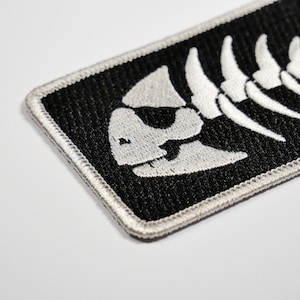May include: White embroidered fish skeleton on a black patch with a white border.