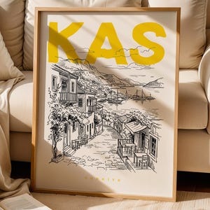 Può includere: Stampa artistica incorniciata con un'illustrazione in bianco e nero di una città costiera, con la parola "KAS" in giallo. La stampa include la parola "TÜRKIYE" in basso. L'opera d'arte è esposta su uno sfondo beige.