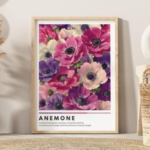 Può includere: Una stampa floreale incorniciata con una vivace composizione di fiori di anemone nei toni del rosa, viola e bianco. La stampa include la parola "ANEMONE" e una descrizione del simbolismo del fiore. L'opera d'arte è esposta su una parete beige.