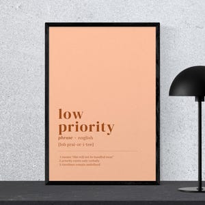 May include: A framed print with the words "low priority" in a brown font on a peach background. The print is in a black frame and is next to a black desk lamp.