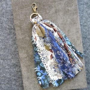 May include: A handmade tassel keychain with a gold-colored clasp and leaf charms. The tassel features fabric strips in shades of blue, white, and patterned floral designs, accented with small brown beads. A unique accessory.