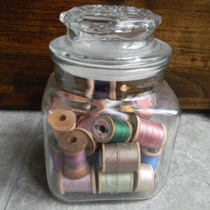 May include: A glass jar with a lid filled with spools of colorful thread. The spools are in shades of pink, blue, green, brown, and white.