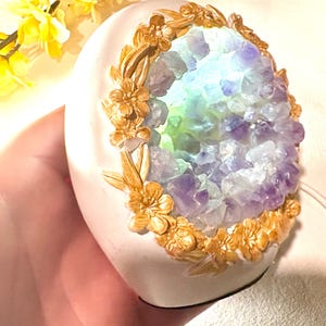 以下が含まれることがあります： 中央に紫と青の結晶の塊がある、白い楕円形の装飾品。結晶は金色の花の模様で囲まれています。オブジェクトは内側から照らされ、柔らかな光を放っています。