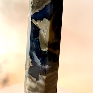 May include: A polished stone tower with a hexagonal shape, featuring a smooth surface with a marbled pattern of dark navy blue, muted olive green, and creamy white tones, ending in a pointed, faceted top with a small translucent white tip.