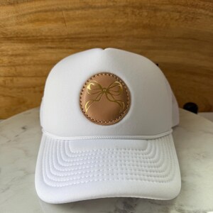 May include: White trucker hat with a curved brim and a mesh back. The hat features a round, tan leather patch with a gold bow design in the center. A white rope detail runs across the front of the hat.