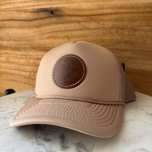 May include: Tan trucker hat with a brown leather patch featuring a floral design. The hat has a curved brim, a tan front panel, and a mesh back. A tan rope detail accents the brim. The hat is sitting on a marble surface.