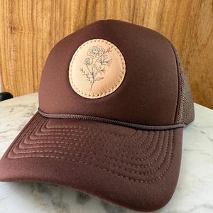 May include: A brown trucker hat with a floral design on a circular patch. The hat features a mesh back, a curved brim, and a brown rope detail. The floral design is a simple line drawing of a flower and leaves.