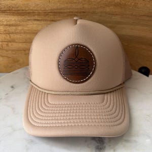 May include: Tan trucker hat with a brown leather patch featuring a graphic design. The hat has a curved brim, mesh back, and a rope detail across the front. The hat is made of a soft, durable material.