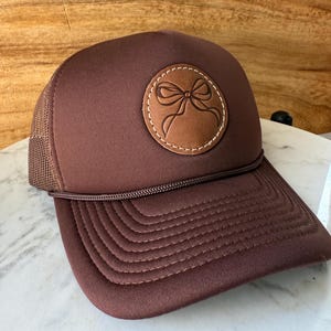 May include: A brown trucker hat with a mesh back and a curved brim. The front of the hat features a circular leather patch with a stitched bow design. The hat has a brown rope detail above the brim.