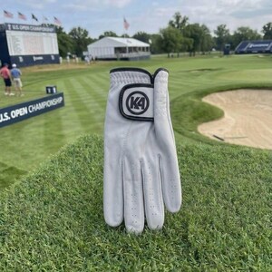 Puede incluir: Un guante de golf gris claro con un acento negro y un logotipo K4. El guante está sobre un campo de golf verde, con un letrero del Campeonato Abierto de EE. UU. en el fondo.