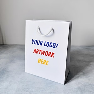 May include: White paper shopping bag with a white rope handle. The bag has the text "YOUR LOGO/ARTWORK HERE" printed in blue, red, and yellow.