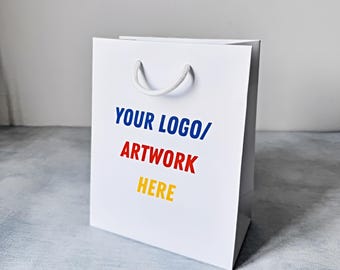 May include: White paper shopping bag with a white rope handle. The bag has the text "YOUR LOGO/ARTWORK HERE" printed in blue, red, and yellow.