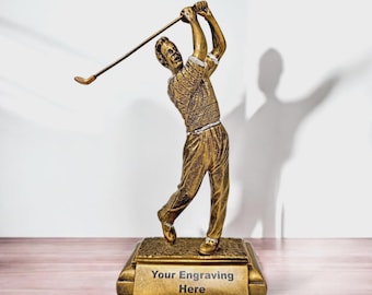 May include: A gold-colored golf trophy depicting a golfer in mid-swing. The figure is on a rectangular base with the text "Your Engraving Here". The trophy is set against a white background and a wooden surface.