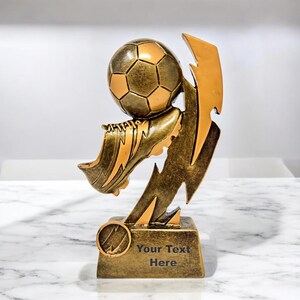 May include: A gold-colored soccer trophy featuring a soccer ball, a soccer cleat, and a lightning bolt design. The base of the trophy has the text "Your Text Here".
