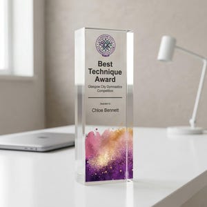 May include: A clear acrylic award with the text "Best Technique Award" and "Glasgow City Gymnastics Competition." The award is rectangular with a colorful design of pink, gold, and purple at the bottom. The name "Chloe Bennett" is also on the award.