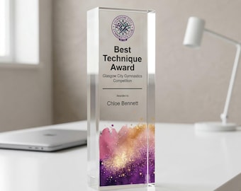 May include: A clear acrylic award with the text "Best Technique Award" and "Glasgow City Gymnastics Competition." The award is rectangular with a colorful design of pink, gold, and purple at the bottom. The name "Chloe Bennett" is also on the award.
