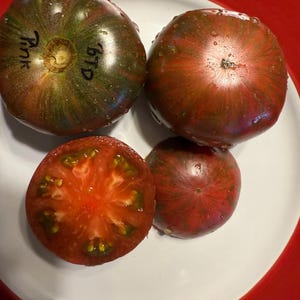 Może przedstawiać: Cztery pomidory dziedzictwa na białym talerzu w czerwonej misce. Dwa całe pomidory są ciemnozielone i czerwone, z napisem "Pink" i "BTD" na jednym. Jeden pomidor jest przekrojony na pół, ukazując czerwone wnętrze.