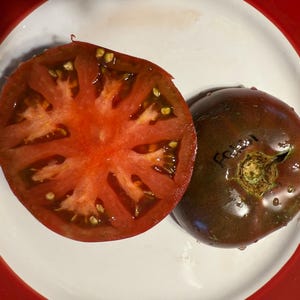 May include: A sliced heirloom tomato with a deep red interior and visible seeds, next to a whole tomato with a dark, almost black, exterior. The tomatoes are on a white plate with a red rim.