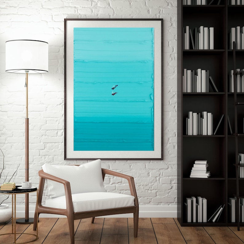 May include: A framed abstract painting with a gradient of blue and turquoise colors. The painting is hung on a white brick wall in a room with a white armchair and a black bookshelf.