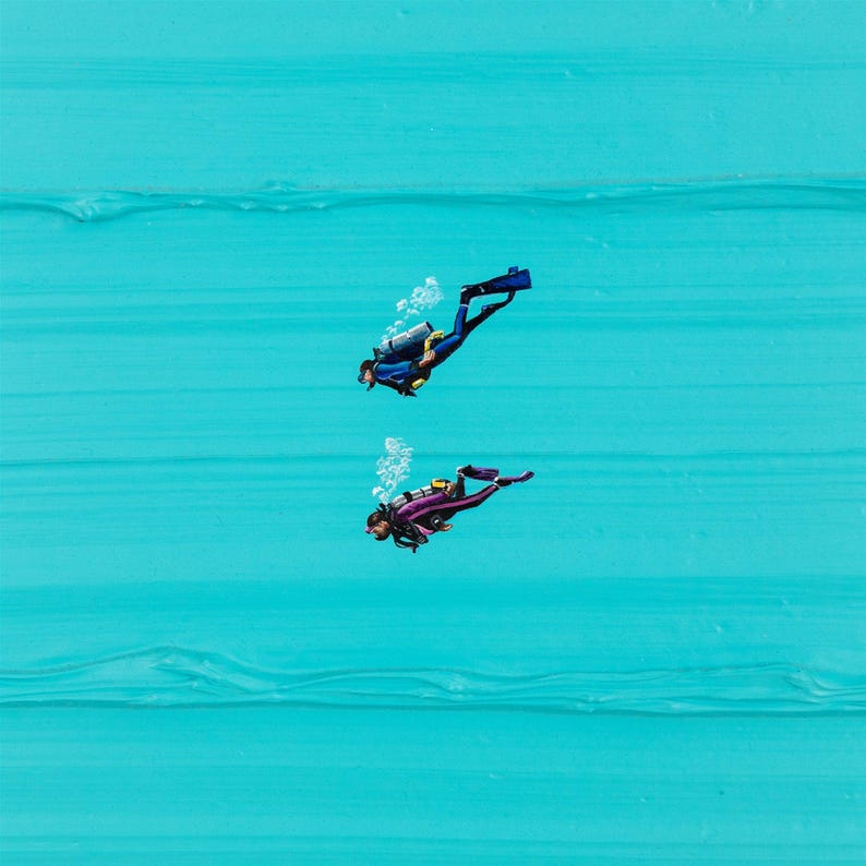 May include: Two scuba divers in blue and purple wetsuits with scuba gear are swimming underwater in a turquoise blue ocean.
