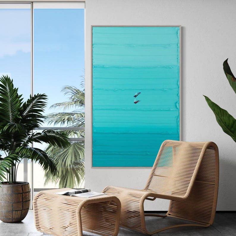 May include: A teal and turquoise abstract painting with horizontal brushstrokes. The painting is framed in a silver frame and hangs on a white wall. A wicker chair and ottoman are in the foreground.