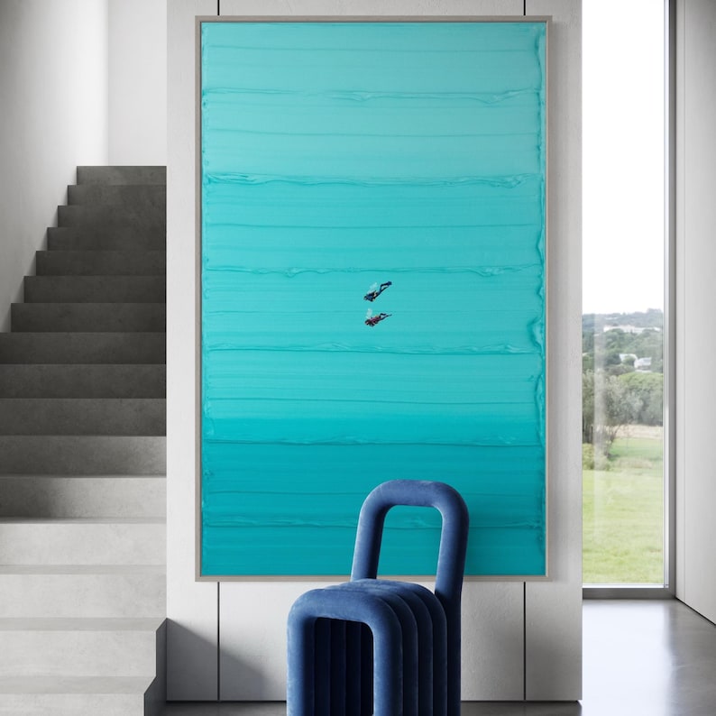 May include: A teal blue abstract painting with horizontal brushstrokes, depicting two figures diving underwater. The painting is framed and hanging on a white wall, with a blue chair in the foreground.