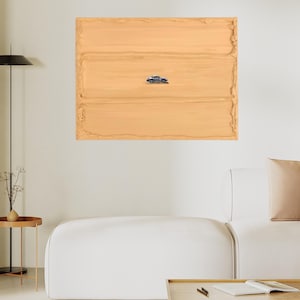 K&ouml;nnte beinhalten: Ein gerahmtes Kunstwerk mit einer kleinen, detaillierten Autoillustration, die sich auf einem strukturierten, hellbraunen Hintergrund befindet. Das Kunstwerk wird &uuml;ber einem wei&szlig;en Sofa und einem Holztisch pr&auml;sentiert, was ein minimalistisches Interieur schafft.
