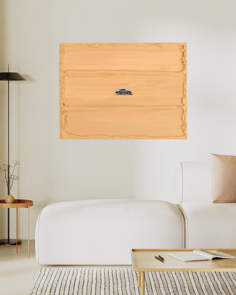 K&ouml;nnte beinhalten: Ein gerahmtes Kunstwerk mit einer kleinen, detaillierten Autoillustration, die sich auf einem strukturierten, hellbraunen Hintergrund befindet. Das Kunstwerk wird &uuml;ber einem wei&szlig;en Sofa und einem Holztisch pr&auml;sentiert, was ein minimalistisches Interieur schafft.