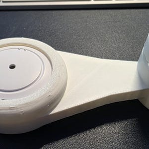 May include: A white 3D-printed device with a circular base and a curved support. The base has a central disc with a small hole. The support has three rectangular cutouts. The device is designed to hold a round object.