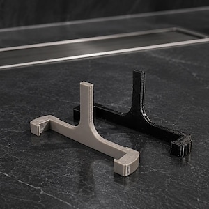 May include: Two T-shaped stands, one in light gray and one in black, featuring a horizontal base with upturned ends and a central vertical support post. These rigid, angular objects are designed with a flat profile and a smooth, uniform finish.