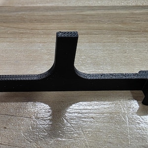 May include: A black, 3D-printed object with a T-shaped design. The object has a horizontal bar with curved ends and a vertical support in the center. The item is likely a holder or stand for an electronic device.