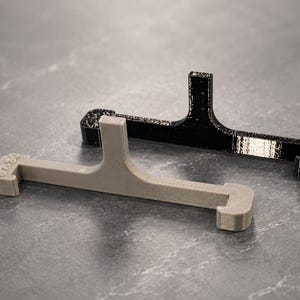 May include: Two T-shaped plastic brackets, one in matte beige and one in black, featuring a horizontal bar with hooked ends and a vertical support post. The beige bracket has the text 67-80 embossed on the top left corner of the horizontal bar.