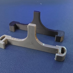 May include: Two plastic, T-shaped objects. One is light gray, the other is dark gray. Both have a rectangular base and a vertical support. The light gray object has a small hole on each end and a small inscription.