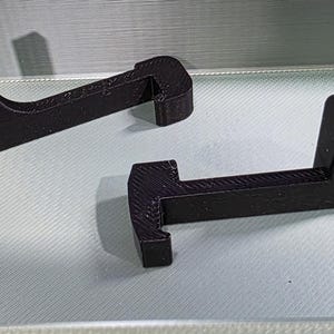 May include: Two black, 3D-printed support brackets. Each bracket has a T-shaped design with a hook on one end and a vertical support. The brackets are resting on a light gray surface.