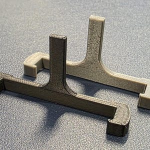 May include: Two 3D-printed, gray and black, plastic stands. Each stand has a rectangular base with a vertical support and a small, rectangular extension. The stands are designed to hold an item.