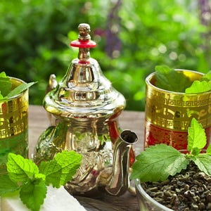 Pode incluir: Uma chaleira prateada com uma parte superior vermelha e dourada, dois copos com acabamentos dourados e uma tigela com folhas de chá verde. Ramos de hortelã fresca estão nos copos e ao redor da chaleira. A cena é montada em uma mesa de madeira com um fundo verde desfocado.