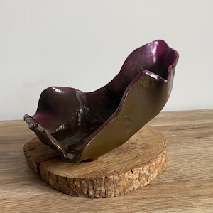May include: A dark purple and brown ceramic bowl with an organic, wavy shape. The bowl is sitting on a wooden base.