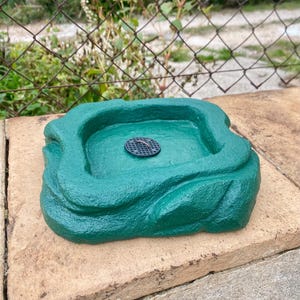 Peut inclure: Un bain d'oiseaux en plastique vert avec une surface texturée et un drain noir au centre. Le bain d'oiseaux est posé sur une surface en pierre brune.