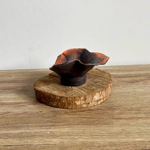 Könnte beinhalten: Eine kleine, schwarze Keramikschale mit einem welligen Rand und einer rot-orangenen Glasur im Inneren. Die Schale steht auf einer Holzscheibe.