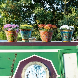 May include: Four decorative flower pots, each filled with colourful flowers. The pots are hand-painted with detailed patterns and sit on a green surface. The background shows foliage and a boat's porthole.