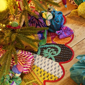 May include: A scalloped, circular tree skirt made from patchwork fabric featuring various colourful patterns, including floral, geometric, and paisley prints in shades of red, yellow, green, and black, with visible white kantha stitching.