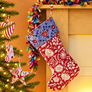 May include: A quilted Christmas stocking featuring a red and white floral pattern on the main body and a contrasting blue and red floral print on the cuff. The stocking includes a fabric loop at the top for hanging and has a soft, padded texture.