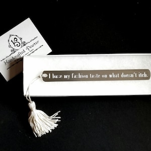 May include: A silver bookmark with a white tassel. The bookmark has the text "I base my fashion taste on what doesn't itch."