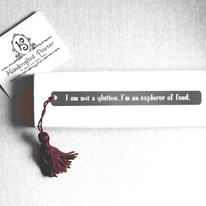 May include: A silver bookmark with a tassel. The bookmark has the text "I am not a glutton. I'm an explorer of food." engraved on it.